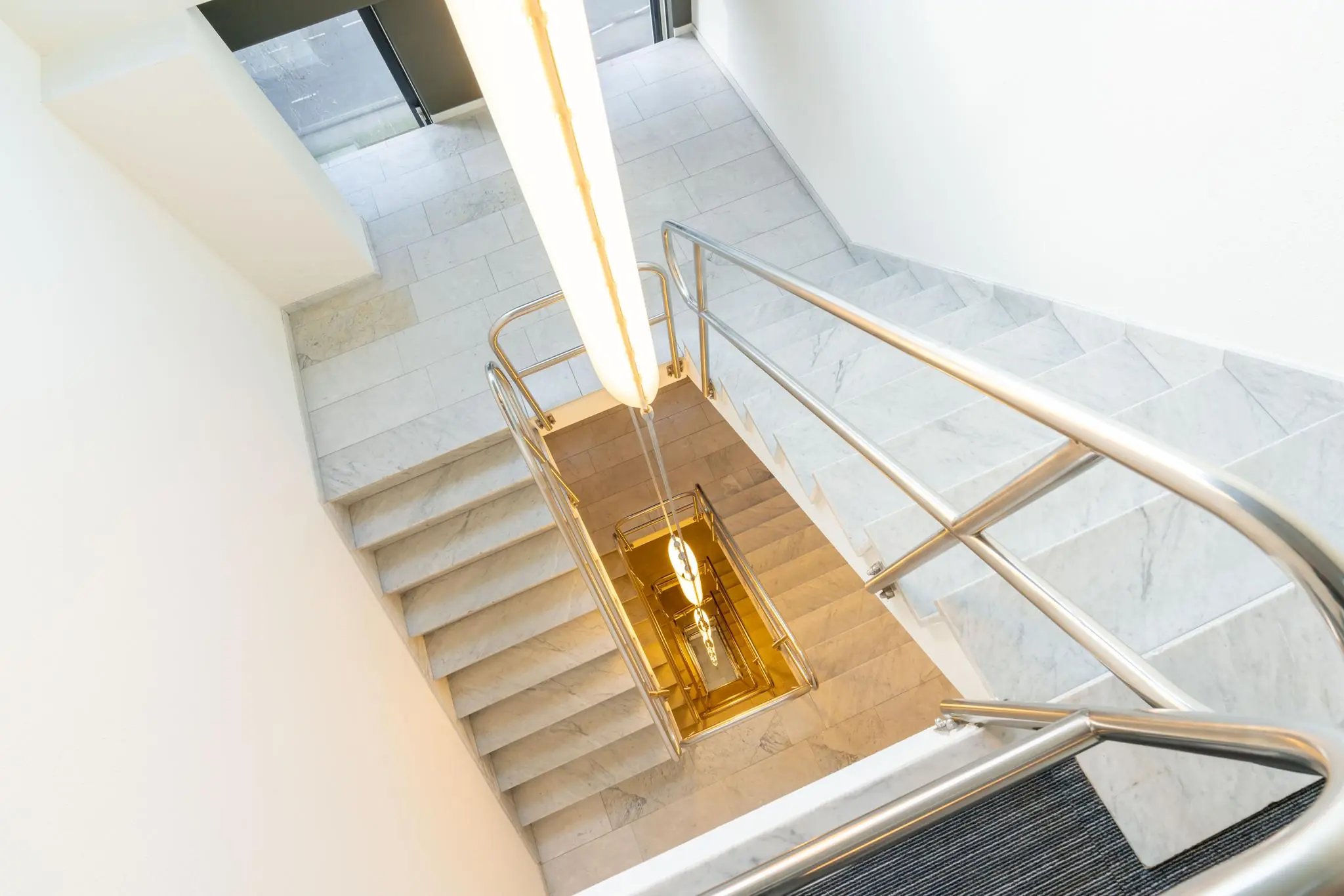 Marmeren trappenhuis met roestvrijstalen leuningen en een opvallende verticale lamp in een gebouw aan de De Bruyn Kopsstraat.